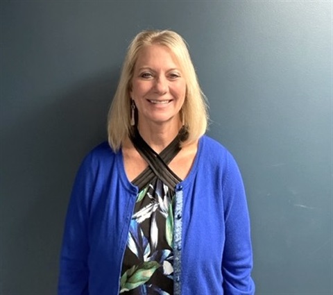 A headshot of a white woman with blond hair down to her shoulders. She's standing up against a blue wall. She's wearing a blue cardigan over a black and white top. 